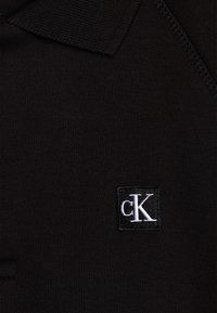 Svart polo-shirt med en texturerad krage. Har en rektangulär logotyp med vit brodering av "cK." Slät tygtextur.