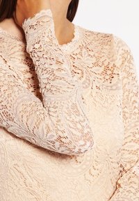 Spitzenlangarmshirt in sanftem Beige mit floralem Muster, gewelltem Ausschnitt und strukturierten Details an den Ärmeln. Leichter, zarter Stoff.