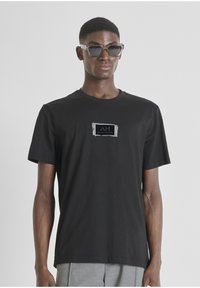 Camiseta de algodón negra con mangas cortas, que cuenta con un parche de logo rectangular en el pecho en colores contrastantes. Textura suave.