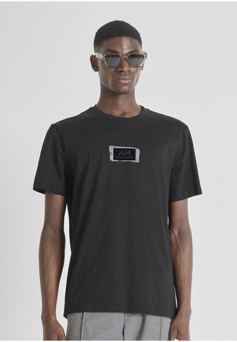 Camiseta de algodón negra con mangas cortas, que cuenta con un parche de logo rectangular en el pecho en colores contrastantes. Textura suave.