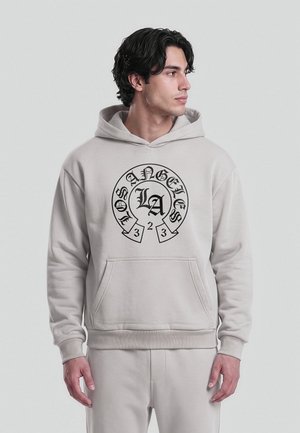 Jeune homme portant un sweat à capuche beige clair et un pantalon, le sweat à capuche affiche un texte noir "Los Angeles LA 323" en motif circulaire sur le devant, regardant sur le côté.