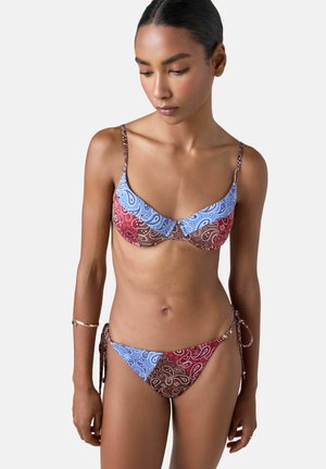 Kvinde iført en bikini med paisley-mønster i blå, rød og brun med justerbare skulderstropper og sidebind, stående foran en ensfarvet baggrund.
