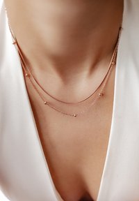 Des colliers en or rose superposés avec des chaînes délicates et de petits accents sphériques, reposant sur une clavicule nue contre un vêtement blanc.