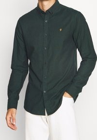 Camicia button-up verde scuro realizzata in un tessuto texturizzato, con colletto a punta e un piccolo logo dorato sul petto, abbinata a pantaloni chiari.