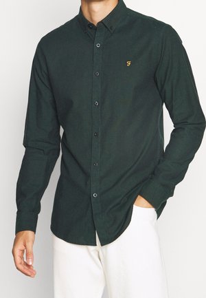 Camisa abotonada verde oscura hecha de una tela texturizada, con un cuello pico y un pequeño logo dorado en el pecho, combinada con pantalones claros.