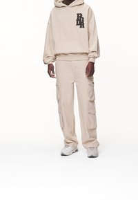 Beige hoodie med "PGRR"-logotyp, stor framficka; matchas med byxor i cargo-stil med sex fickor och avslappnad passform. Lätta sneakers.