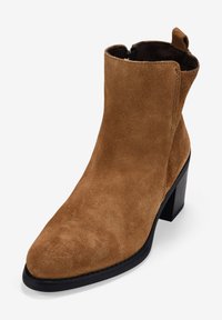 Bota de tobillo de ante marrón con puntera puntiaguda y tacón cuadrado negro. Presenta una tira en la parte superior para facilitar su colocación.