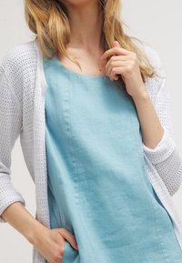 Abito in lino azzurro chiaro con scollo rotondo, abbinato a un cardigan bianco lavorato a maglia con vestibilità ampia e maniche a tre quarti.