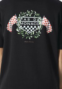 Sort T-shirt med et grafisk design med en laurbærkrans, ternede flag og teksten "PAS DE MONACO" i hvidt og rødt.