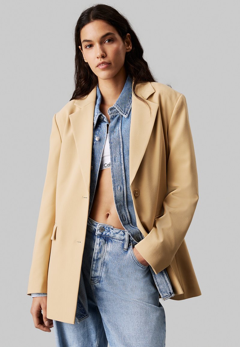 Beżowy dopasowany żakiet z wyraźnymi ramionami, nałożony na dżinsową kurtkę i białą bluzkę, stylizowany z jasnoniebieskimi jeansami o wysokim stanie.