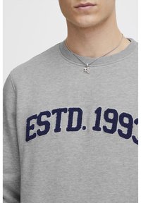 Grå hettegenser med marineblå brodert tekst "ESTD. 1993" på forsiden. Har en klassisk rund hals og myk tekstur. Sølvkjede er synlig.