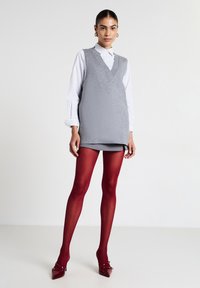 Maglione grigio senza maniche sopra una camicia bianca a righe, abbinato a collant rossi profondi e décolleté a punta. Texture liscia e design a strati.