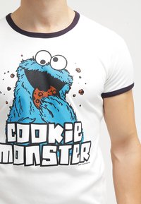 Weißes T-Shirt mit einem blauen Cookie Monster Grafik, das ein lächelndes Gesicht und einen Keks in der Hand zeigt. Schwarze und blaue Buchstaben darunter. Rundhalsausschnitt.