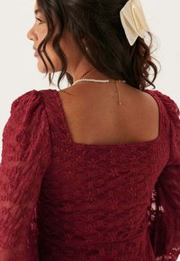 Robe en dentelle bordeaux avec un décolleté carré, des manches bouffantes et un motif floral texturé, accompagnée d'un collier en perles et d'un léger accessoire en forme de nœud.