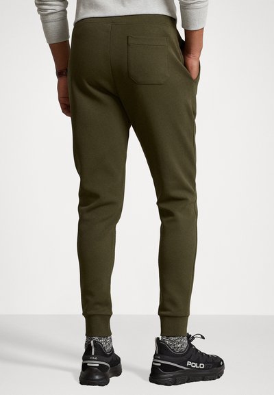 Slim-fit oliwkowe dresy wykonane z miękkiego materiału, z tylną kieszenią i ściągaczami na kostkach, w połączeniu z czarnymi trampkami i wzorzystymi skarpetkami.