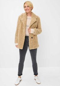 Cappotto in pile beige con collo a punte, indossato sopra un maglione leggero, abbinato a jeans scuri e sneakers bianche. Il cappotto ha un aspetto testurizzato.