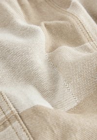 Beige und cremefarbener Textilstoff mit einer strukturierten Oberfläche und einem gewebten Muster, das diagonale Streifen und sichtbare Nähte aufweist. Weiche, fließende Optik.