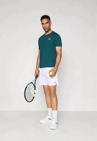 Τιρκουάζ κοντομάνικο αθλητικό μπλουζάκι, λευκές βερμούδες, κρατώντας μια μπάλα τένις και ρακέτα, με λευκά αθλητικά παπούτσια και κάλτσες μέχρι τον αστράγαλο. Λογότυπο στο μπλουζάκι.