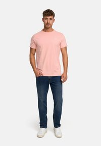 Light pink cotton t-shirt with short sleeves, round neckline, paired with dark blue jeans and white sneakers, standing on a plain background.