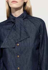 Navy blue button-up shirt with a high collar and large bow detail, featuring a textured fabric and gold-tone buttons.