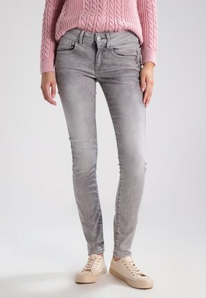 Person trägt hellgraue Slim-Fit-Jeans, einen rosa kabelgestrickten Pullover und beige Sneaker und steht vor einem schlichten hellen Hintergrund.