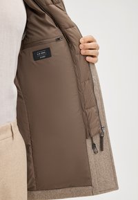 Płaszcz z recyklingowanej wełny z brązową, pikowaną podszewką. Posiada kieszeń na zamek i metkę. Zewnętrzny materiał ma teksturowany wzór rybiej ości.