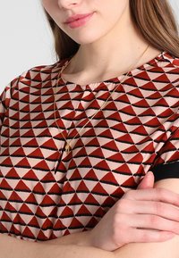 Rotes und pinkes gemustertes Oberteil mit dreieckigen Formen, rundem Ausschnitt und kurzen schwarzen Ärmeln; geschichtete goldene Halskette mit Anhänger.