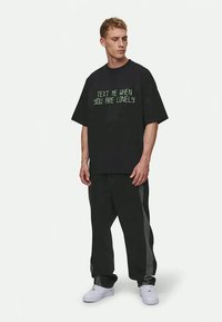 Črna boste pametno tshirt z zelenim besedilom "PIŠITE MI, KO STE OSAMLJENI." Nosite z ohlapnimi črnimi hlačami in belimi supergami. Preprost dizajn.