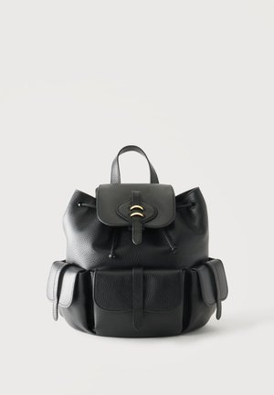 Sac à dos en cuir noir avec rabat frontal et trois poches extérieures sur un fond clair uni.