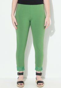 Leggings verdes con una cintura elástica, con un acento de encaje en el dobladillo. Llevados con sandalias negras sobre un fondo claro.