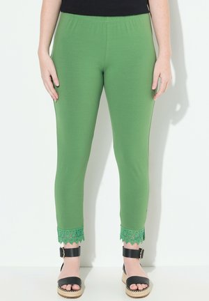 Leggings verts avec une taille élastique, présentant une accent en dentelle à l'ourlet. Portés avec des sandales noires sur un fond clair.