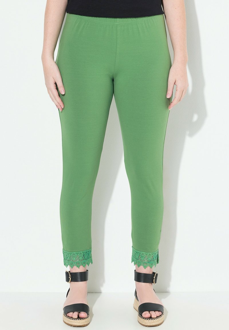 Leggings verdes con una cintura elástica, con un acento de encaje en el dobladillo. Llevados con sandalias negras sobre un fondo claro.