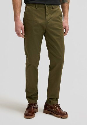 Homme portant un pantalon droit vert olive et des chaussures en cuir marron à lacets, debout sur un fond clair uni.