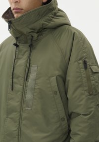 Olivno zelena podložena jakna z visokim ovratnikom, nastavljivo kapuco, sprednjimi žepi in žepom na rokavu z zadrgo. Narejena iz gladkega, trpežnega materiala.