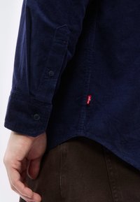 Gros plan sur la main d'une personne près du poignet d'une chemise en velours côtelé bleu marine avec des boutons noirs et le logo Levi's rouge, au-dessus d'un pantalon marron foncé.