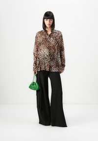 Mulher vestindo uma blusa de estampado leopardo castanho, calças largas pretas, segurando uma pequena bolsa verde, em pé contra um fundo branco.
