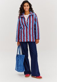 Prošívaná bunda s modro-vínovým pruhovaným vzorem, s patentkami, doplněná modrou semišovou taškou, rozšířenými tmavými džíny a červenými botami.
