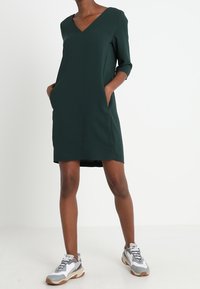 Grünes Kleid mit V-Ausschnitt, dreiviertel Ärmeln und Seitentaschen. Getragen mit grauen und weißen Sportschuhen. Glatte Stoffstruktur.