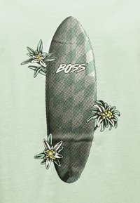 Σανίδα του σερφ με ένα πουά καρό μοτίβο και τη λέξη "BOSS", περιτριγυρισμένη από τρία λευκά και κίτρινα άνθη εδέλβαϊς σε ανοιχτό πράσινο φόντο.