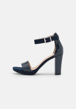 Sandal en cuir verni bleu marine à talons hauts avec une bride de cheville, bout ouvert, semelle plateforme et un talon épais. La bride est ornée d'une boucle en argent.