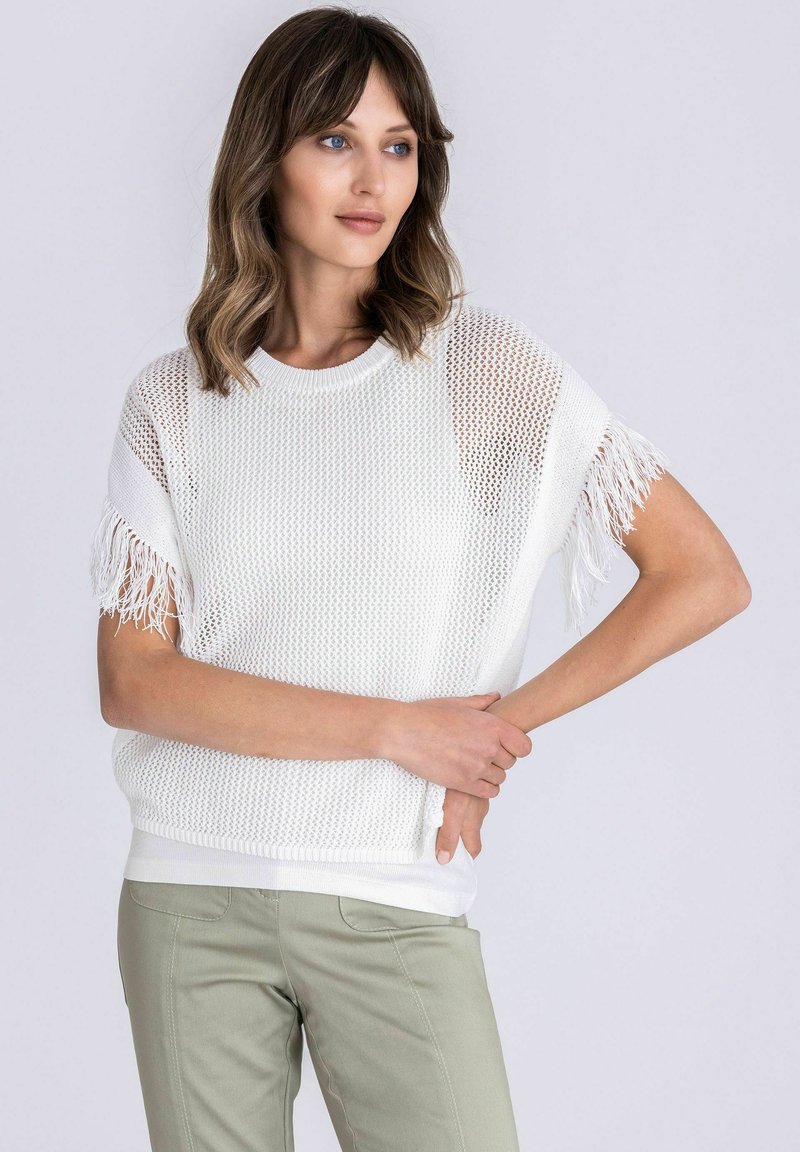 Vrouw met schouderlang haar, gekleed in een witte gebreide top met franjes aan de mouwen en lichtgroene broek, staand met gekruiste armen tegen een witte achtergrond.