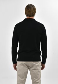 Zwarte polo met lange mouwen en kraag, gemaakt van gebreid materiaal. In combinatie met lichtgekleurde broeken, gezien vanaf de achterkant.