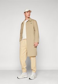 Beige trenchcoat over een witte kabelgebreide trui, gecombineerd met crèmekleurige joggers en witte sneakers. Bevat het Lacoste-logo op de jas.