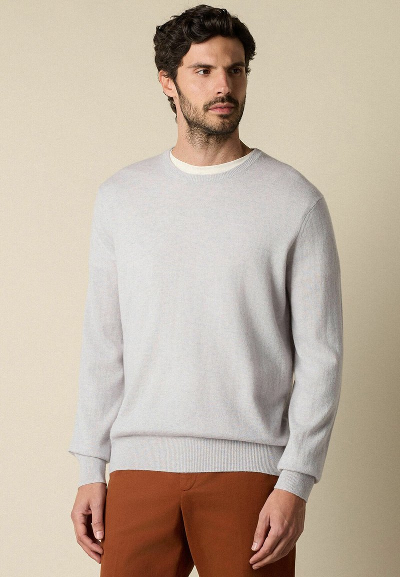 Svetle šedý pletený svetr s kulatým výstřihem, ribbovaným lemem a dlouhými rukávy, spárovaný s hnědými kalhotami. Měkká textura a volný střih.