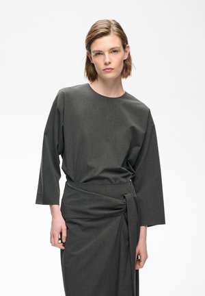 Jeune personne aux cheveux courts portant une robe gris foncé à manches longues avec un détail noué à la taille sur fond blanc.