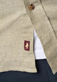 Lys beige linned skjorte med krave, træknap og et lille burgunder mærke med en silhuetdesign nær kanten.