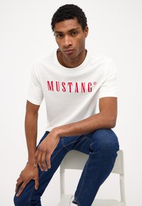 Balta medvilninė marškinėlių su raudonu "MUSTANG" užrašu, trumpomis rankovėmis, apvaliu iškirpimu, derinami su mėlyniais džinsais, sėdintys ant kėdės.