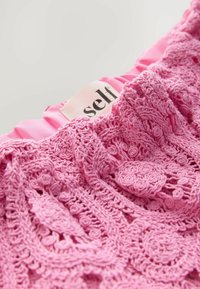Tessuto all'uncinetto rosa con un motivo floreale testurizzato e arricciature in satin, caratterizzato da un'etichetta che recita "Self." Design morbido e dettagli intricati.