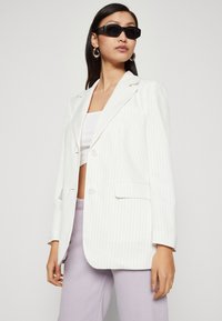 Blazer bianco a righe con vestibilità rilassata, chiusura con un solo bottone, rever a punta e tasche laterali, abbinato a un top corto e pantaloni lavanda.