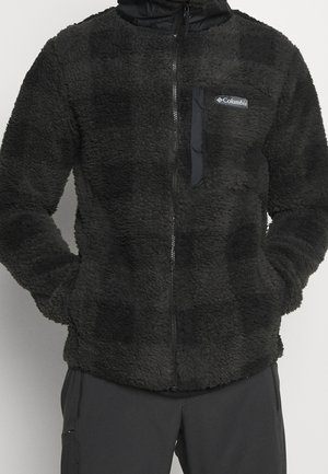 Musta fleecetakki, jossa edessä vetoketju, rintatasku ja Columbian logo, käytetty mustien housujen kanssa, kädet sivutaskuissa.